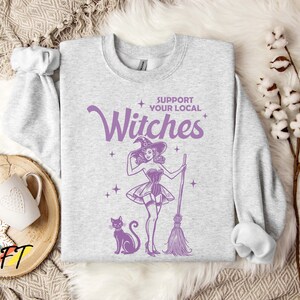 May include: A grey sweatshirt with the text "Support Your Local Witches" in purple lettering. A cartoon witch with a black cat and a broom is featured on the sweatshirt.