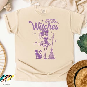 May include: A beige t-shirt with a purple graphic of a witch holding a broom and a black cat. The text "Support Your Local Witches" is printed in purple on the shirt.