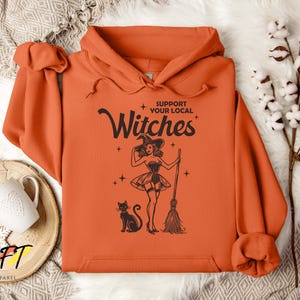 May include: Orange hoodie with a black graphic of a witch holding a broom and a black cat. The text on the graphic says "Support Your Local Witches".