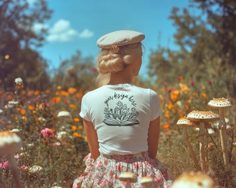 Può includere: Una maglietta bianca con un design floreale sul retro. Il design presenta un libro con fiori che crescono da esso. Il testo "your design here" è scritto sopra il libro. La maglietta è indossata da una persona in un campo di fiori.