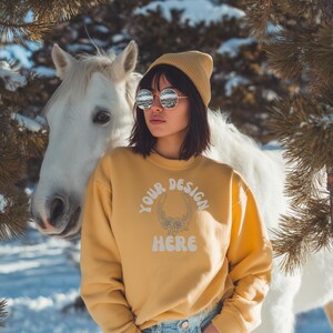 Könnte beinhalten: Eine Person trägt ein gelbes Sweatshirt, eine Mütze und eine runde Sonnenbrille vor einem weißen Pferd. Auf dem Sweatshirt steht der Text "YOUR DESIGN HERE" in einem kreisförmigen Design. Der Hintergrund umfasst Schnee und Nadelbaumzweige.