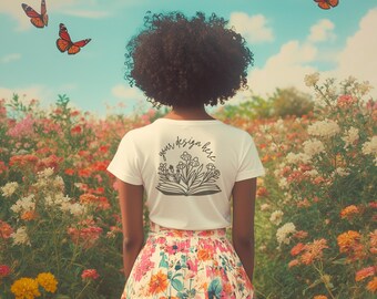Può includere: Una maglietta bianca con un disegno floreale e il testo "your design here" sul retro. La persona che indossa la maglietta è in piedi in un campo di fiori.
