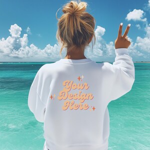 Op de afbeelding: Wit sweatshirt met de tekst "Your Design Here" in een retro lettertype met oranje en roze accenten. Het sweatshirt wordt gedragen door een persoon die voor een blauwe zee en lucht staat.