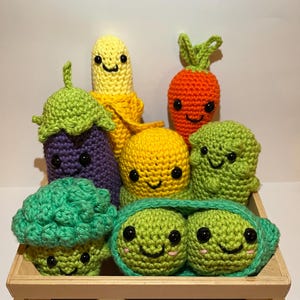 May include: A collection of crocheted plush vegetables in a wooden crate. The vegetables include a purple aubergine, a yellow corn, a yellow pea pod, a green pea pod, a green broccoli, a green pea pod with two peas inside, and an orange carrot.