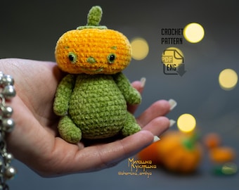 May include: A crocheted pumpkin amigurumi toy with a green body and an orange head. The toy has a friendly face with black eyes and a small mouth. The toy is being held in a hand with a silver bracelet.