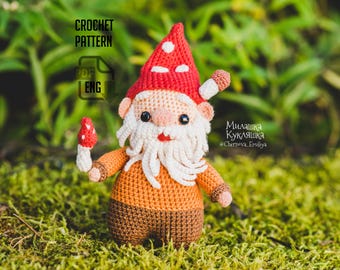May include: A crochet gnome doll featuring a red pointed hat with white spots, a long white beard, a peach-colored face, an orange shirt, and brown pants. The gnome holds a small red mushroom in one hand. Text reads CROCHET PATTERN PDF ENG.