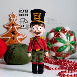 May include: Crochet pattern for a toy nutcracker soldier. The soldier is wearing a red and green uniform with a black hat and a gold belt. The pattern is available in PDF format and is in English.