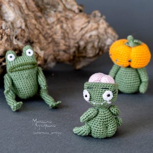 May include: Three crocheted Halloween toys: a green frog, a green zombie with a pink brain, and a green pumpkin with an orange face.
