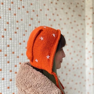 May include: A bright orange knitted hat with white star embroidery. The hat has a long tie that hangs down the back of the neck.