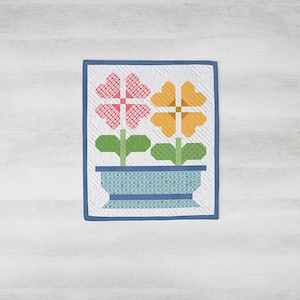 May include: A quilted wall hanging featuring two stylized flowers in a blue patterned pot. One flower is pink, the other is yellow, both with green stems and leaves. The background is white with a blue border.