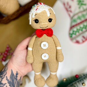May include: A crocheted gingerbread man doll with a white icing-like head and colorful sprinkles. It has a red bow tie, white button details, and white trim on the arms and legs. The doll is tan and has black eyes and a stitched smile.