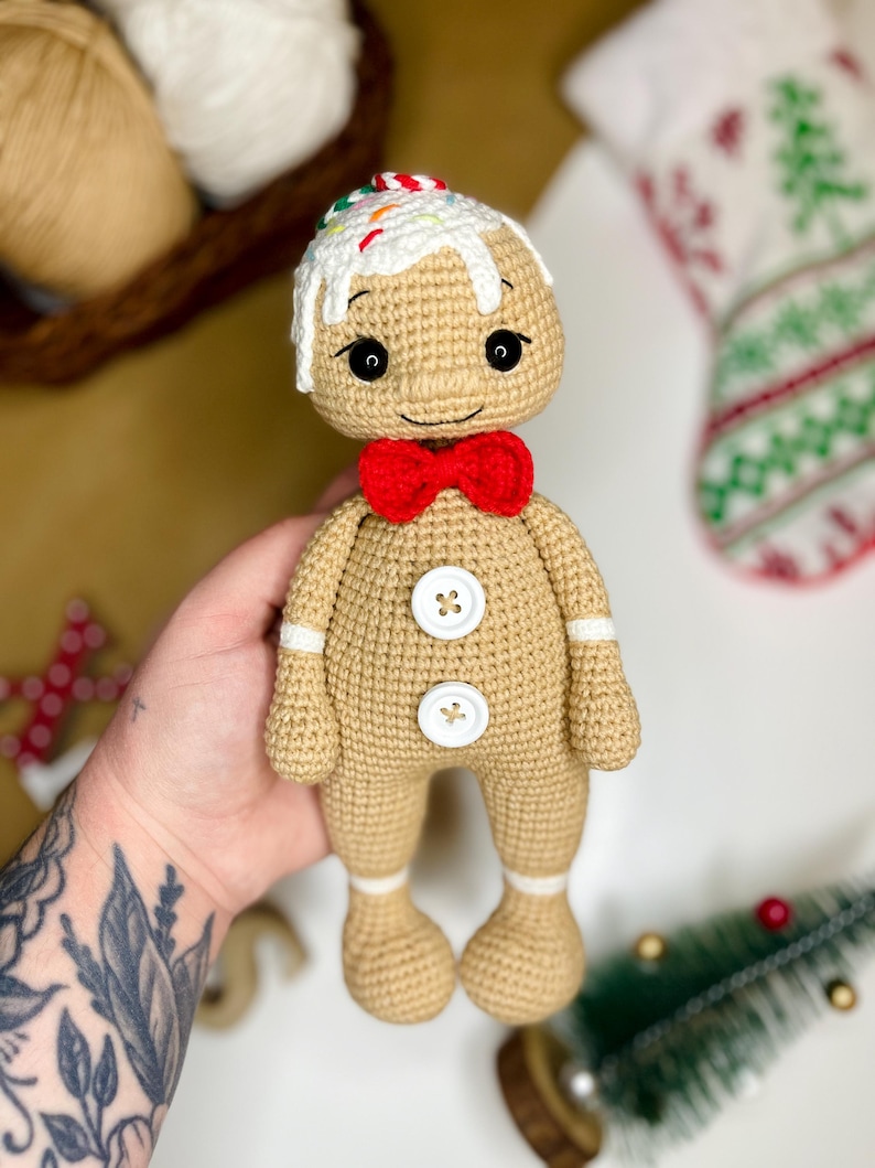 May include: A crocheted gingerbread man doll with a white icing-like head and colorful sprinkles. It has a red bow tie, white button details, and white trim on the arms and legs. The doll is tan and has black eyes and a stitched smile.