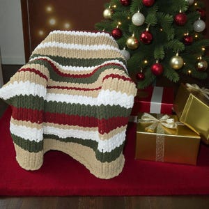 May include: A chunky knit blanket in a chair shape, featuring horizontal stripes of cream, white, green, red, and tan. The blanket is on a red rug, with a decorated Christmas tree and wrapped gifts in the background.