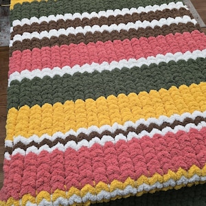 May include: A handmade, colorful blanket with alternating horizontal stripes. The blanket features stripes of pink, yellow, white, brown, and green. The blanket is made of a soft, textured material.