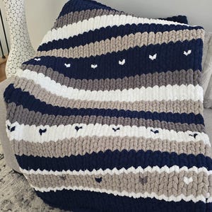 May include: A chunky knit blanket in navy blue, white, and gray stripes, with small white heart-shaped accents. The blanket is draped over a gray sofa, offering a cozy and textured aesthetic. A decorative item for the home.