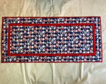 May include: A red, white, and blue quilted table runner featuring a pattern of gnomes wearing red, white, and blue hats. The runner has a red and blue border.