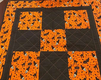 May include: A square quilted table topper featuring a repeating pattern of black cats, skulls, and jack-o'-lanterns on an orange background, interspersed with solid black quilted squares. The fabric has a stitched diamond pattern throughout.