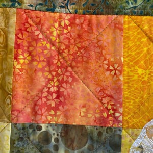 May include: A patchwork quilt featuring a central square block with a vibrant orange and red floral batik pattern. The quilt is constructed with diagonal stitching lines and includes adjacent blocks in shades of yellow, gold, and olive green.