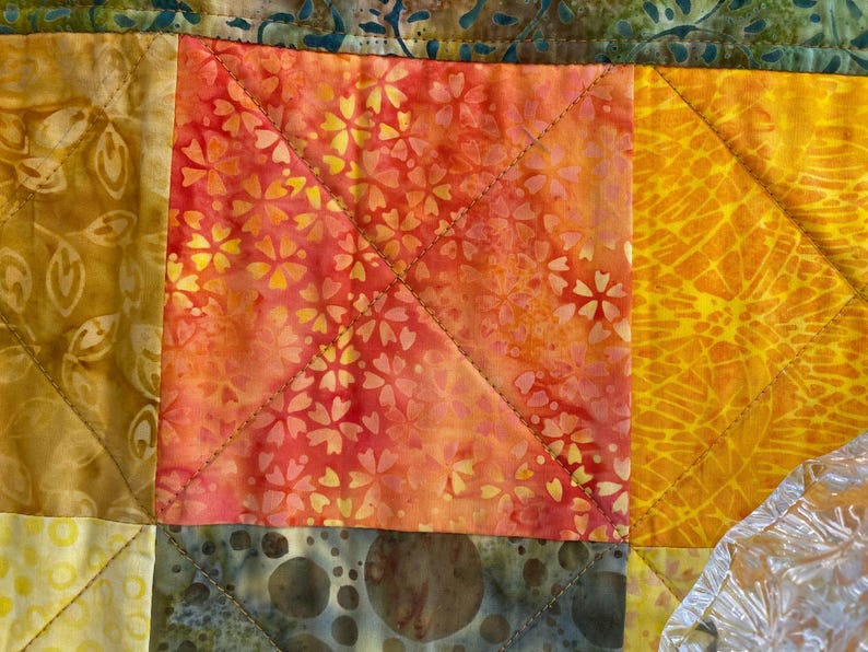May include: A patchwork quilt featuring a central square block with a vibrant orange and red floral batik pattern. The quilt is constructed with diagonal stitching lines and includes adjacent blocks in shades of yellow, gold, and olive green.