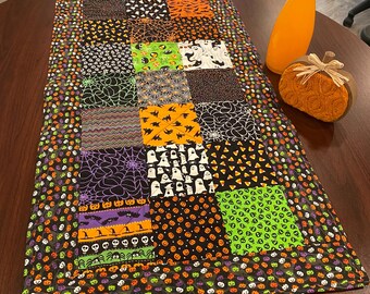 May include: A rectangular patchwork table runner featuring a variety of Halloween-themed fabric squares in black, orange, purple, and lime green. The patterns include ghosts, spider webs, pumpkins, skulls, bats, and candy corn, finished with a border.