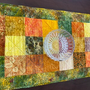 May include: A rectangular quilted table runner featuring a patchwork of fabric squares in shades of yellow, orange, brown, and green with various floral and abstract patterns, topped with a clear, round, faceted glass bowl with a sunburst design.
