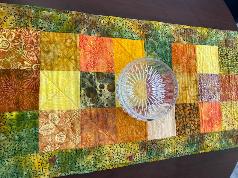 May include: A rectangular quilted table runner featuring a patchwork of fabric squares in shades of yellow, orange, brown, and green with various floral and abstract patterns, topped with a clear, round, faceted glass bowl with a sunburst design.