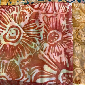 May include: A quilted fabric textile featuring a patchwork design with square blocks. The central block displays a coral and white floral batik pattern, while adjacent sections show golden-brown leaf motifs and teal green patterns with visible stitching.