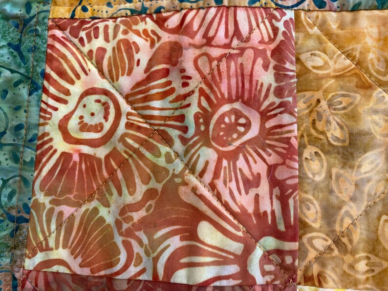 May include: A quilted fabric textile featuring a patchwork design with square blocks. The central block displays a coral and white floral batik pattern, while adjacent sections show golden-brown leaf motifs and teal green patterns with visible stitching.
