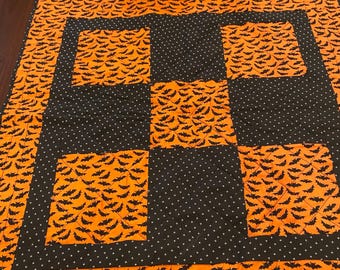 May include: A square quilted table topper featuring a patchwork design of orange fabric with a black bat pattern alternating with black fabric featuring a small white star print. The quilt has a border of orange fabric with black bats.