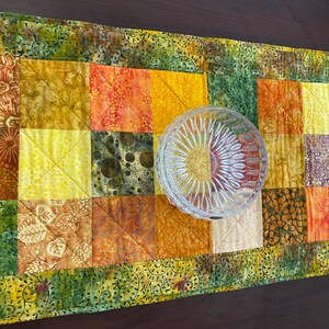 May include: A rectangular quilted table runner featuring a patchwork design of various fabric squares in shades of yellow, orange, and green with floral and abstract patterns, topped with a clear, round, faceted glass bowl with a sunburst design.