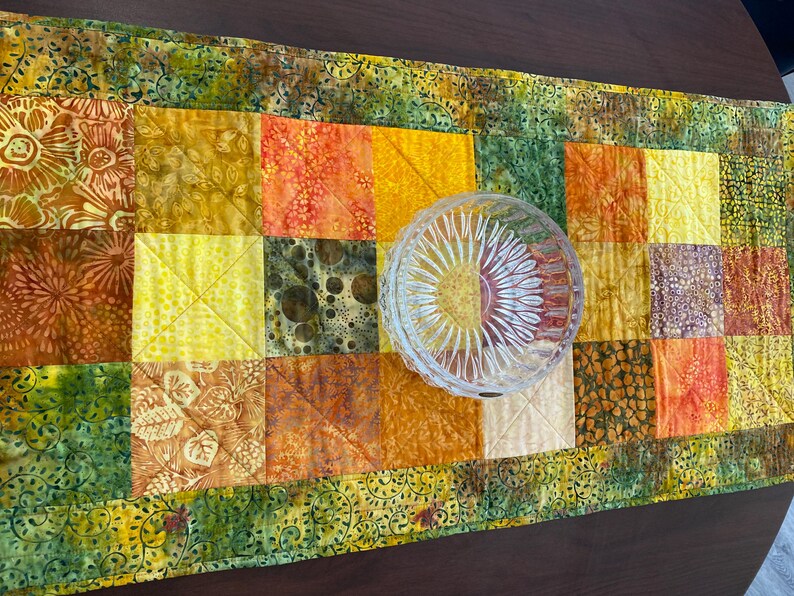 May include: A rectangular quilted table runner featuring a patchwork design of various fabric squares in shades of yellow, orange, and green with floral and abstract patterns, topped with a clear, round, faceted glass bowl with a sunburst design.