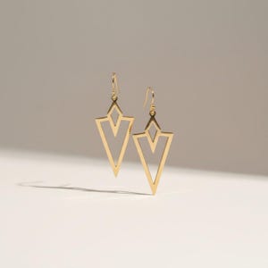 May include: A pair of gold-toned drop earrings featuring a geometric, open-frame arrowhead design. Each earring is suspended from a simple hook wire and consists of a large triangular outline with a smaller, nested triangular cutout inside.