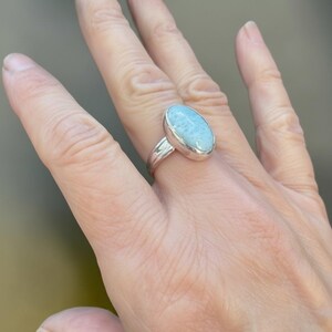 May include: A silver ring featuring a smooth, oval-shaped, light blue Larimar gemstone set in a polished metal bezel. The stone displays natural white marbling patterns across its surface, and the band is a simple, solid silver metal construction.