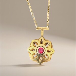 May include: A gold-colored pendant necklace featuring a floral design with a central red gemstone. The pendant is suspended from a delicate gold chain. The necklace is a piece of jewelry.