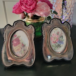 May include: Two silver picture frames with ornate edges, each displaying a floral print. The frames are sitting on a black surface.