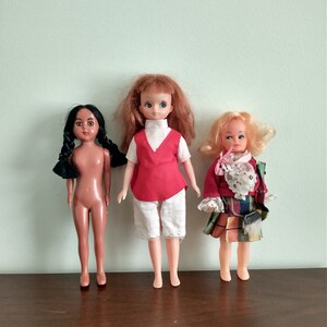 Puede incluir: Tres muñecas vintage con diferentes peinados y atuendos. La muñeca de la izquierda tiene el pelo negro y un cuerpo desnudo. La muñeca del centro tiene el pelo castaño y lleva un chaleco rojo, pantalones cortos blancos y calcetines blancos. La muñeca de la derecha tiene el pelo rubio y lleva una falda de cuadros rojos y verdes con un cuello de encaje blanco.