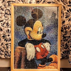 以下が含まれることがあります： ミッキーマウスのカラフルなモザイク画像が描かれた額縁付きジグソーパズル。ミッキーは茶色の丸太に座っており、トレードマークの赤いショートパンツと白い手袋を着用しています。パズルは青い背景に配置されています。