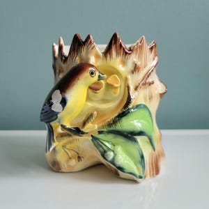 Könnte beinhalten: Ein Keramik-Pflanztopf in Form eines Baumstumpfes mit einem gelben Vogel, der auf einem Ast sitzt. Der Vogel hat braune und weiße Markierungen und schaut nach rechts. Der Topf ist mit grünen Blättern und brauner Rinde verziert.