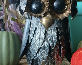May include: A black metal owl with gold accents and large black eyes. The owl is standing on a woven surface.