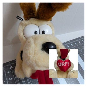 May include: A plush dog toy with large white eyes, a black nose, and floppy brown ears. The dog has a red tongue and a small inset image of the dog holding a red heart with the word "URF!" on it.