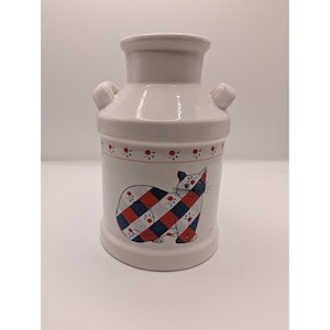 May include: White ceramic milk jug with a cat design. The cat is patterned with red and blue checks. The jug has a small handle on each side and a red floral pattern around the top.
