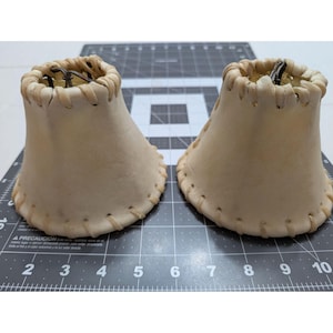 May include: Two off-white leather lamp shades with a stitched edge and a flared base. The shades have a bell shape and are designed to fit over a light bulb. The top edge is also stitched.