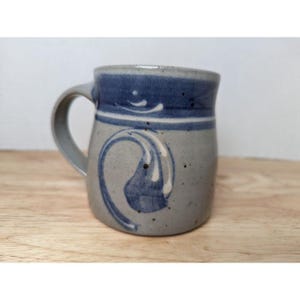 May include: A handmade ceramic mug with a gray base and a blue rim and decorative swirl. The mug has a curved handle and is set against a light wooden surface.