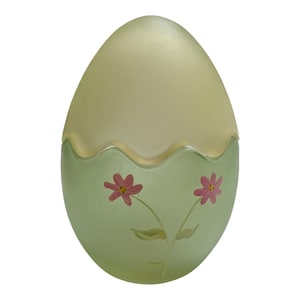 May include: An egg-shaped decorative container with a light green base and a pale yellow top. The base features two pink flowers with green stems and leaves. The container has a frosted glass appearance.