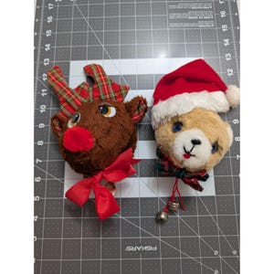 May include: Two Christmas ornaments: a brown reindeer head with plaid antlers and a red bow, and a teddy bear head wearing a Santa hat. The bear has blue eyes and a green and red plaid bow. Both have a festive holiday theme.