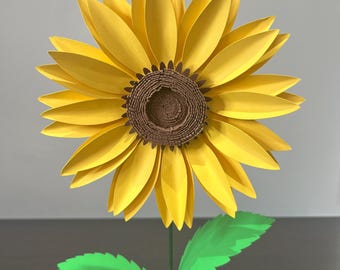 May include: A paper craft sunflower with yellow petals and a brown center. The sunflower has green leaves and a green stem.