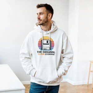 Puede incluir: Sudadera con capucha blanca con un diseño retro de disquete sobre un fondo de puesta de sol. El texto dice "THE ORIGINAL CLOUD STORAGE". La sudadera tiene una capucha con cordón y un bolsillo delantero.