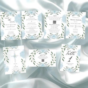 May include: A collection of wedding stationery featuring a soft blue floral design. The set includes invitations, RSVP cards, and table cards. Each card has a watercolor floral wreath and text, including "Save the Date" and "Table 1".