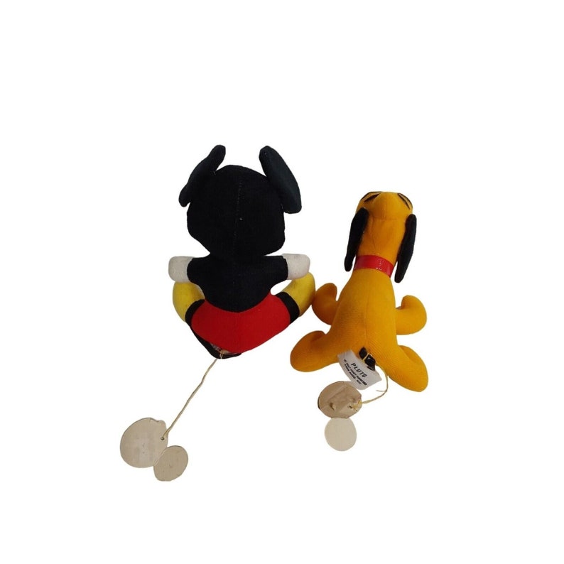 Op de afbeelding: Twee kleine pluche Disney-personagespeeltjes. E&eacute;n is Mickey Mouse, met een zwart lichaam, een rode broek en gele schoenen. De andere is Pluto, een gele hond met zwarte oren en een rode halsband. Beide hebben touwtjes en labels.