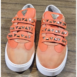 May include: Pair of orange sneakers with three hook-and-loop straps adorned with silver triangle studs. The sneakers have white soles and a suede-like toe. The tongue of the shoe has the text "Valentina Rangoni Firenze".